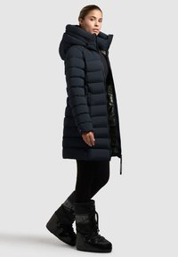 Mujer con un abrigo de plumas negro largo y con capucha, leggings negros y botines de invierno negros, de pie contra un fondo claro y liso.