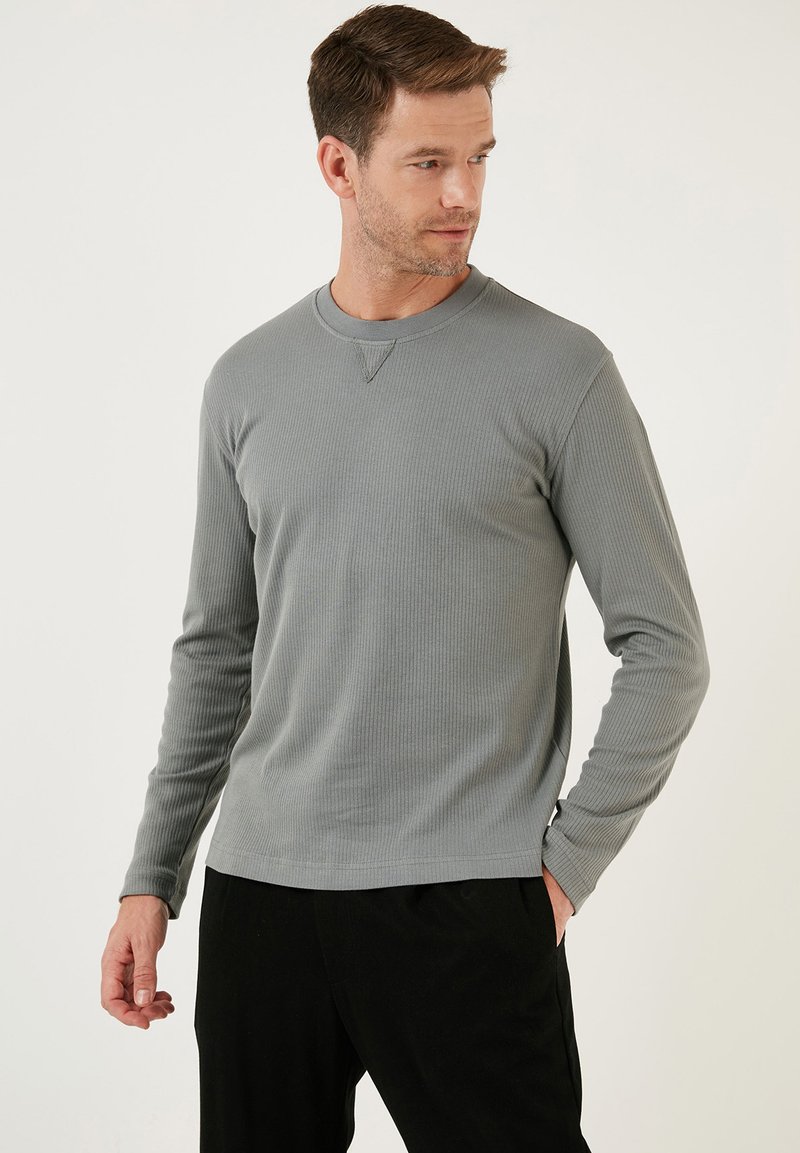 Világosszürke bordás, hosszú ujjú póló kerek nyakkivágással és diszkrét V-öltéssel. Fekete nadrággal viselve. Puha tapintású és testhezálló fazonú.