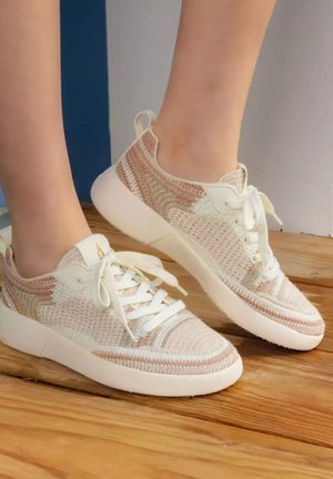 Baskets en tricot beige et blanc avec des lacets blancs portées aux pieds, debout sur un sol en bois avec un fond neutre.