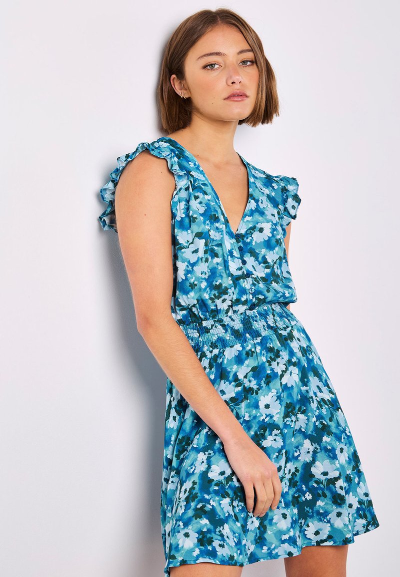 Blaues Blumenkleid mit tiefem V-Ausschnitt, Rüschennähten an den Ärmelchen und elastischem Taillenbund. Der Stoff zeigt verschiedene Blautöne und weiße Blumen.