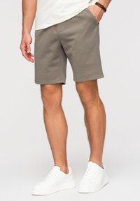 Homme portant un short beige longueur genou et des baskets blanches, debout avec une main dans la poche sur un fond clair uni.