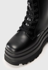 Botas de combate de cuero negro y brillante con una suela gruesa, punta redonda y cordones negros gruesos, que presentan un pequeño logotipo en el lateral.