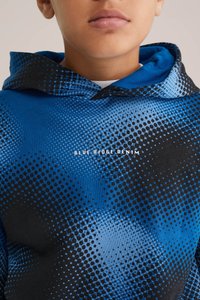 Hooded sweatshirt ar zilu un melnu punktu rakstu, izgatavots no mīksta auduma. Uz krūtīm ir uzraksts "BLUE RIDGE DENIM."