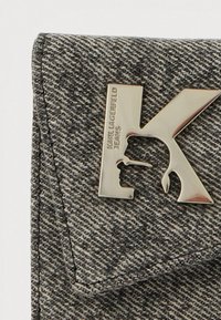 Pelēks džinsa maks ar teksturētu virsmu, uz kura izceļas metālisks "K" logotips un iegriezts dizainera vārds papildus detaļām.