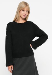 Pulôver preto com um corte descontraído, mangas longas e bainha canelada. Apresenta tachas decorativas nos ombros. Textura suave.