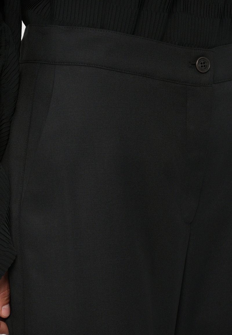 Détail de pantalons noirs ajustés avec fermeture à boutons et poches latérales, portés avec un haut noir texturé.