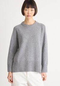 Frau trägt einen schlichten grauen Wollpullover mit langen Ärmeln und gerippten Bündchen, kombiniert mit weißen Hosen, steht vor einem weißen Hintergrund.