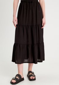 Falda larga negra con volantes, hecha de un tejido ligero, que presenta una cinturilla elástica y una silueta fluida. Combinada con sandalias negras con tachuelas.