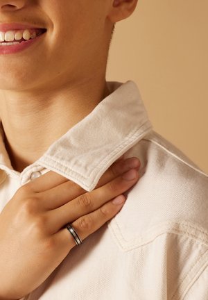 Witte coll shirt met een textuurstof. Hand rust op de kraag, die een zilveren ring met een zwarte streep toont. Zachte, neutrale achtergrond.