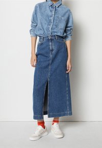 Jeansskjortan har en krage och en framficka. En lång jeanskjol med en framklippning och fransad nederkant matchas med vita sneakers och orange strumpor.