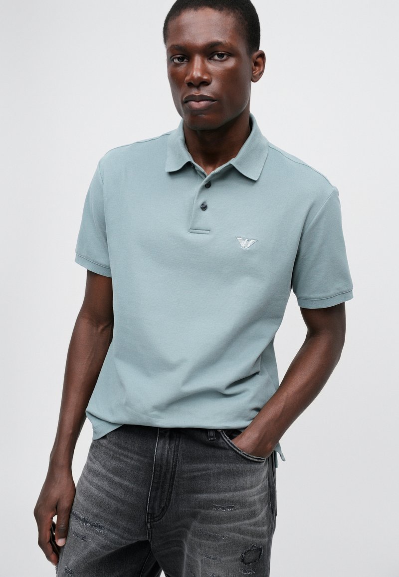 Polo shirt bleu clair avec un col, trois boutons noirs et un petit logo brodé sur la poitrine gauche ; porté avec un jean gris délavé.