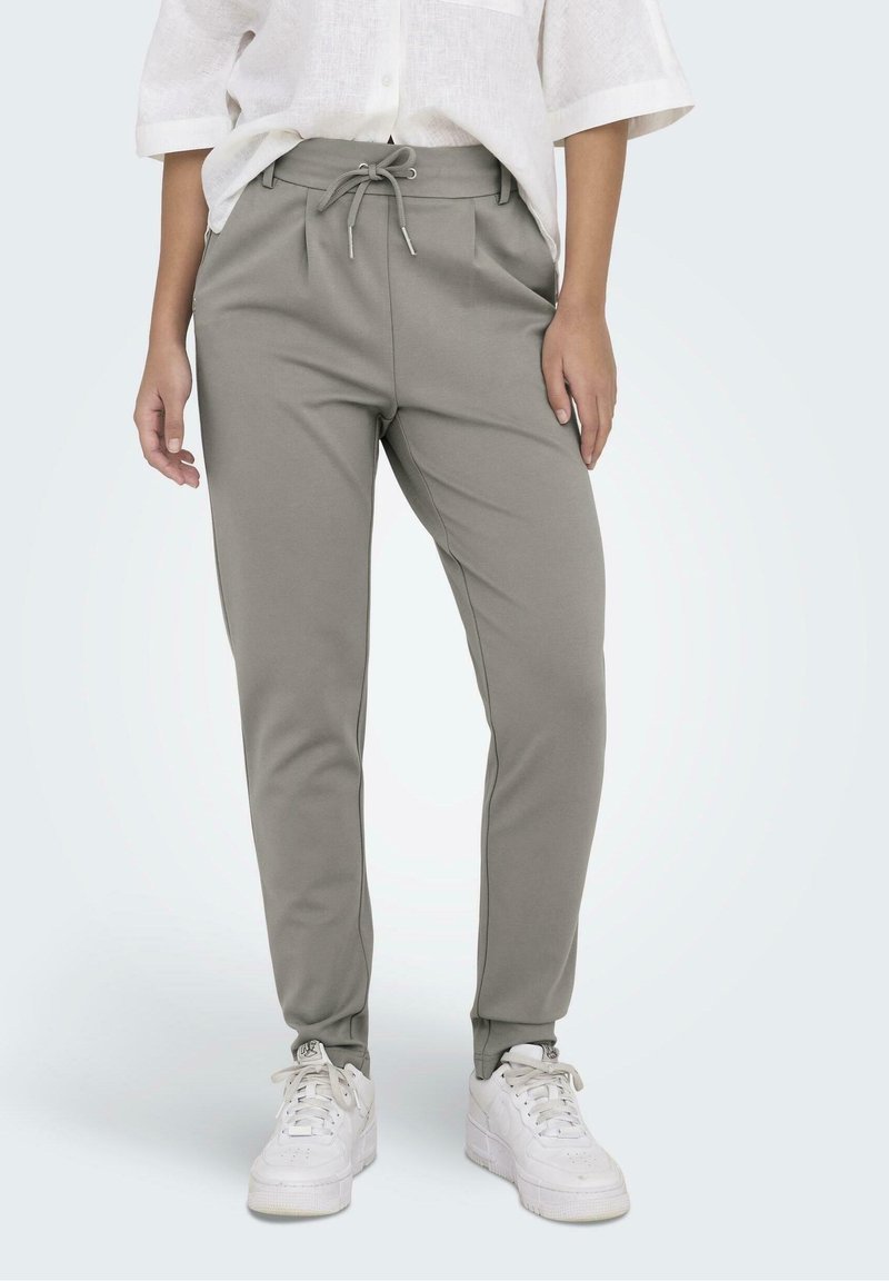 Personne portant un pantalon gris clair fuselé avec cordon, des baskets blanches et une chemise blanche à boutons avec les manches retroussées.