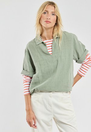 Chemise en lin vert clair avec un col en V, manches retroussées, portée avec un haut à rayures rouges et blanches en dessous. Associée à un pantalon blanc.