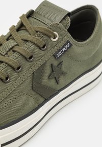 Sneaker in tela verde con suola in gomma, caratterizzata da un logo a stella nera e dettagli in tessuto, lacci rotondi e un'etichetta con logo in rilievo sulla linguetta.