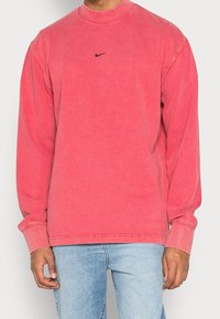 Dlouhý rukáv svetr ve vybledlé červené barvě, z bavlněné směsi. Obsahuje kulatý výstřih a černé logo Nike umístěné na hrudi. Volný střih.