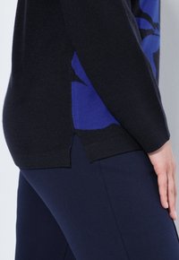 Maglione lavorato a maglia con tessuto nero, motivo floreale blu, fessure laterali all'orlo, abbinato a pantaloni blu scuro, texture liscia.