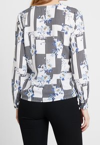 Blusa a maniche lunghe bianca, con una miscela di righe nere e motivi floreali in blu e giallo sul retro. Tessuto di texture liscia.