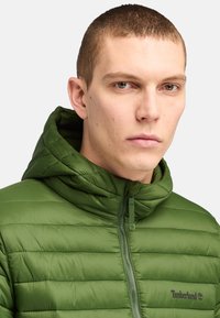Groene pufferjack met capuchon, met een gebold ontwerp, hoge kraag en een rits aan de voorkant. Timberland-logo op de borst.