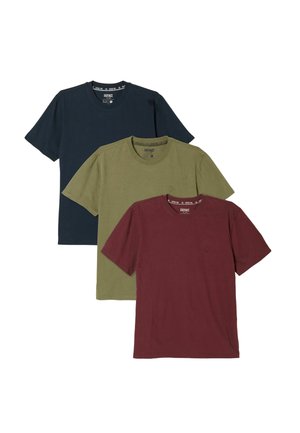 Lächelnder Mann mit Bart und lockigem Haar, der ein dunkelblaues T-Shirt und dunkelblaue Jeans trägt, steht mit den Händen hinter dem Rücken.