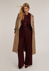 Beige trenchcoat över en vinröd kostym med vida byxor. Har en knapp-uppläggning på toppen och spetsiga klackar, vilket framhäver rena linjer och texturer.