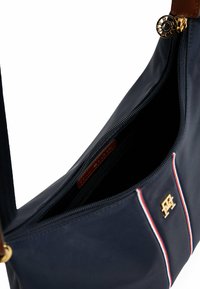 Marineblå nylon skuldertaske med et gyldent logo, cremefarvede og røde striber, med en lynlåsåbning og en brynfarvet læderrem.