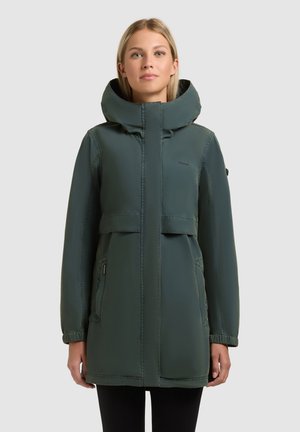 Femme portant un parka d'hiver à capuche vert foncé avec des poches latérales zippées, debout devant un fond clair uni.