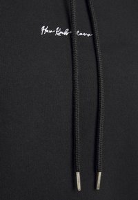 Sudadera negra con cordones trenzados y extremos metálicos. Presenta texto bordado en el pecho. Tejido suave con un diseño ajustado.
