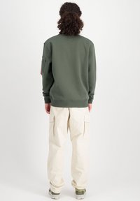 Sudadera verde con cuello redondo y puños de canalé, combinada con pantalones cargo beiges. Detalle notable: bolsillo en la manga izquierda.