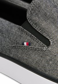 Primer plano de zapato sin cordones texturizado gris con panel elástico lateral negro y pequeña etiqueta de tela roja, blanca y azul sobre la suela blanca.