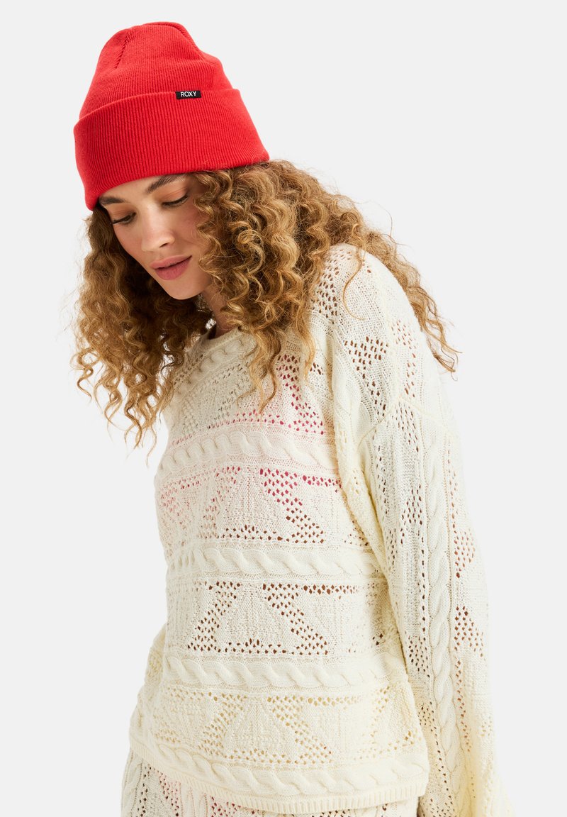 Gebreide trui in crème met open patronen en kabeldetails. Gecombineerd met een felrode geribbelde beanie met een zwart logo.
