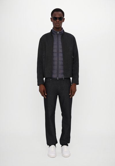 Schwarze Zip-Up-Jacke über einer grauen gepolsterten Weste, kombiniert mit dunklen Jeans und weißen Sneakern. Das Modell trägt eine schwarze Sonnenbrille. Einfaches Design.