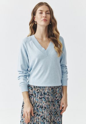 Pulover lejer de culoare albastru deschis, cu decolteu în V și mâneci lungi, având detalii de cusătură subtile. Asortat cu o fustă plisată cu model floral pentru un contrast.