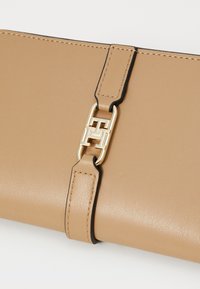 Portefeuille en cuir tan avec bord noir, doté d'un fermoir en métal doré et d'une surface texturée. Design élégant avec un détail de logo central.