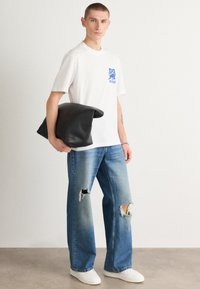 Maglietta bianca con logo blu, indossata con jeans chiari strappati e distorti, abbinata a una borsa a tracolla nera e sneakers bianche.