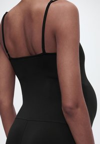 Έγκυος, φοράει εφαρμοστό μαύρο αμάνικο τοπ, φωτογραφημένη από πίσω σε απλό φόντο.