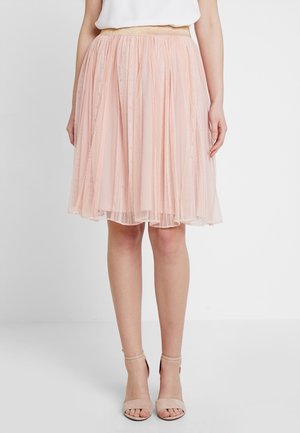 Kvinde iført en lys pink knælang tylskørt med blonde detaljer og et guld elastisk taljebånd, parret med nøgne ankelremssandaler.