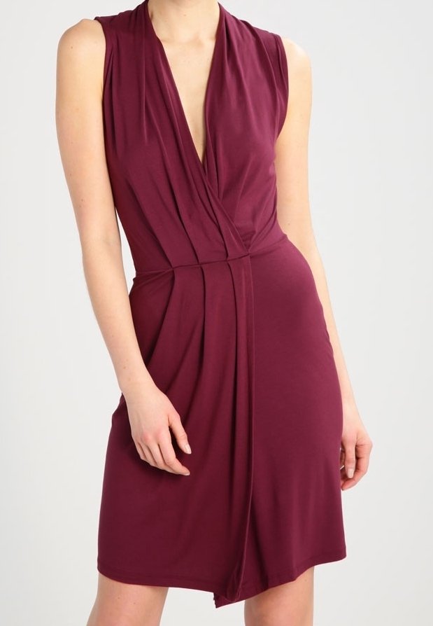 Robe bordeaux sans manches, longueur genou, avec un décolleté en V profond et un design portefeuille plissé portée par une personne debout.
