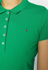 Vihreä polo-paita, joka on valmistettu tekstuurista kankaasta, jossa on kolme napinläpeä ja pieni logo punaisilla, valkoisilla ja sinisillä yksityiskohdilla rinnassa.