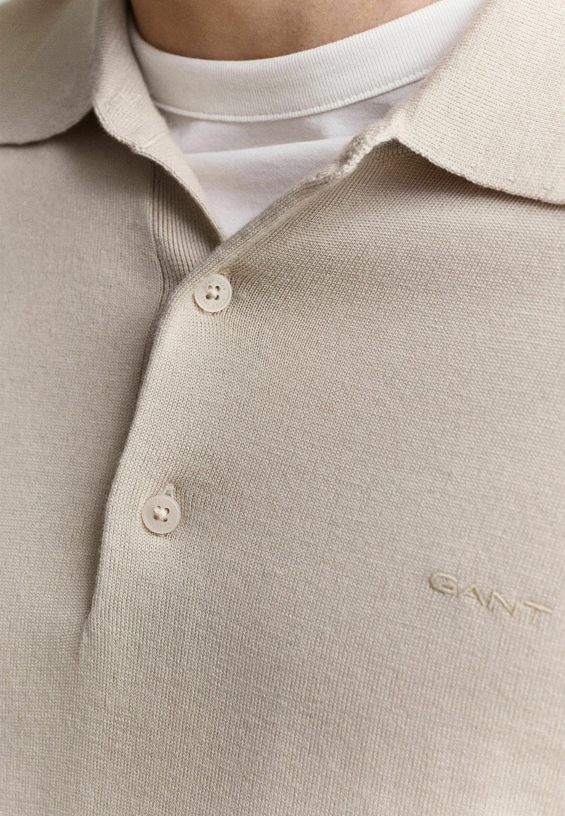 Close-up of beige knit polo shirt with buttons and white undershirt visible, showing subtle embroidered logo on chest.