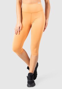 Leggings capri de couleur pêche en tissu extensible, avec une taille haute et une texture lisse. Ensemble porté avec des chaussures de sport noires.