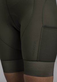 Dark green cycling shorts with padded seat, smooth texture, tight fit, and logo on side. Features contrasting ribbed band at leg openings.