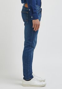 Jeans de ganga skinny fit azuis com desbotamento subtil, design de cinco bolsos, patch de pele na cintura, usados com ténis brancos.