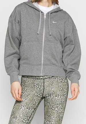 Osoba oblečená v šedé mikině Nike na zip a legínách s vysokým pasem a leopardím potiskem, stojící před jednobarevným pozadím.