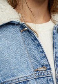 Vaaleansininen farkkutakki, jossa on vaaleancreme Sherpa-niska, näkyvät ompeleet ja ribbattu valkoinen neule alla.