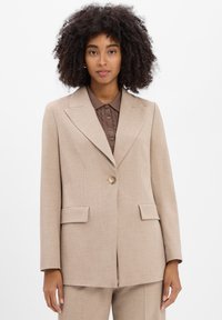 Vrouw met krullend haar, gekleed in een beige colbert over een bruine gebreide overhemd met kraag, staande tegen een effen lichte achtergrond.