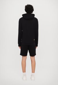 Black sweatshirt with a hood, paired with black shorts. Features ribbed cuffs and a small logo accent on the lower back. Model facing away.