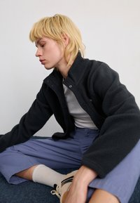 Black fleece jacket with a high collar and snap button closure, paired with light purple pants and cream socks. Textured fabric is visible.