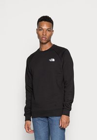 Sudadera negra hecha de material suave con cuello redondo y puños acanalados. Presenta un logo blanco en el lado izquierdo del pecho. Lleva unos jeans azules.