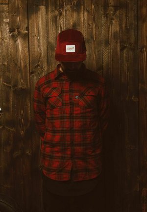 Red plaid shirt with two chest pockets, worn under a red corduroy cap. Background features dark wooden planks. Low-light setting.