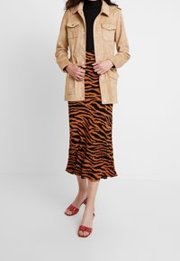 Giacca in suede beige con quattro tasche, dolcevita nero a costine, gonna midi a righe arancioni e nere, e sandali rossi con cinturini.